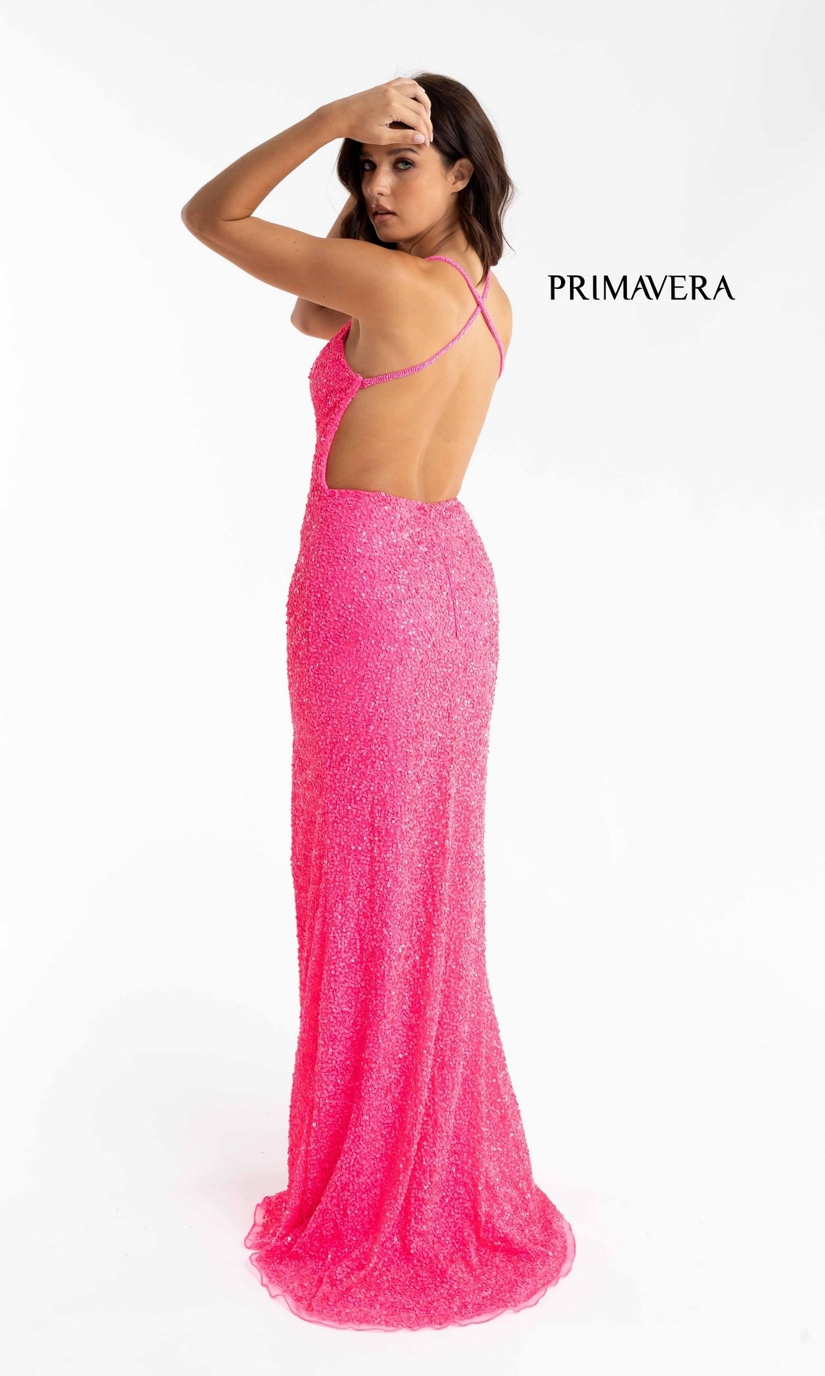 Woman in a sparkly, bright pink Open-Back Long Sequin Prom Dress 3291B with thin straps and a deep V-neck, looks over her shoulder against a plain white background. The word “PRIMAVERA” appears beside her.